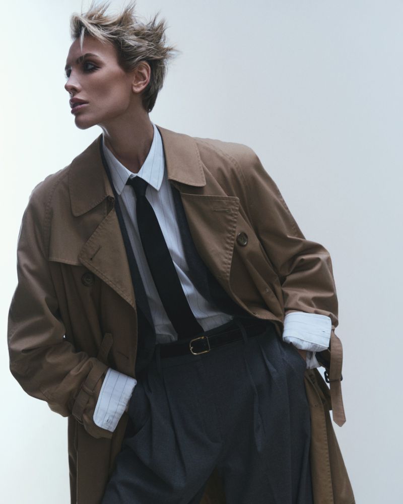 A stylish adult woman in a tan trench coat, white collared shirt, and dark tie poses in a neutral studio setting, fashion-forward and elegant.
