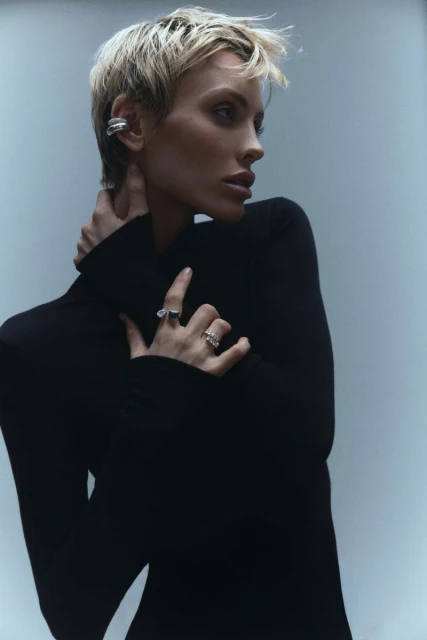 Close-up studio shot of a blonde woman with short hair wearing an elegant black turtleneck dress and silver rings and ear cuff.