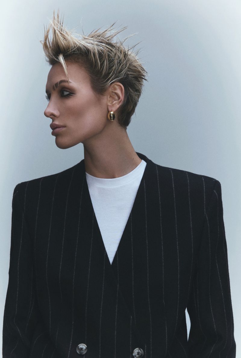 Wallis Day with spiky blonde hair wearing a black pinstripe blazer and white t-shirt against a grey background