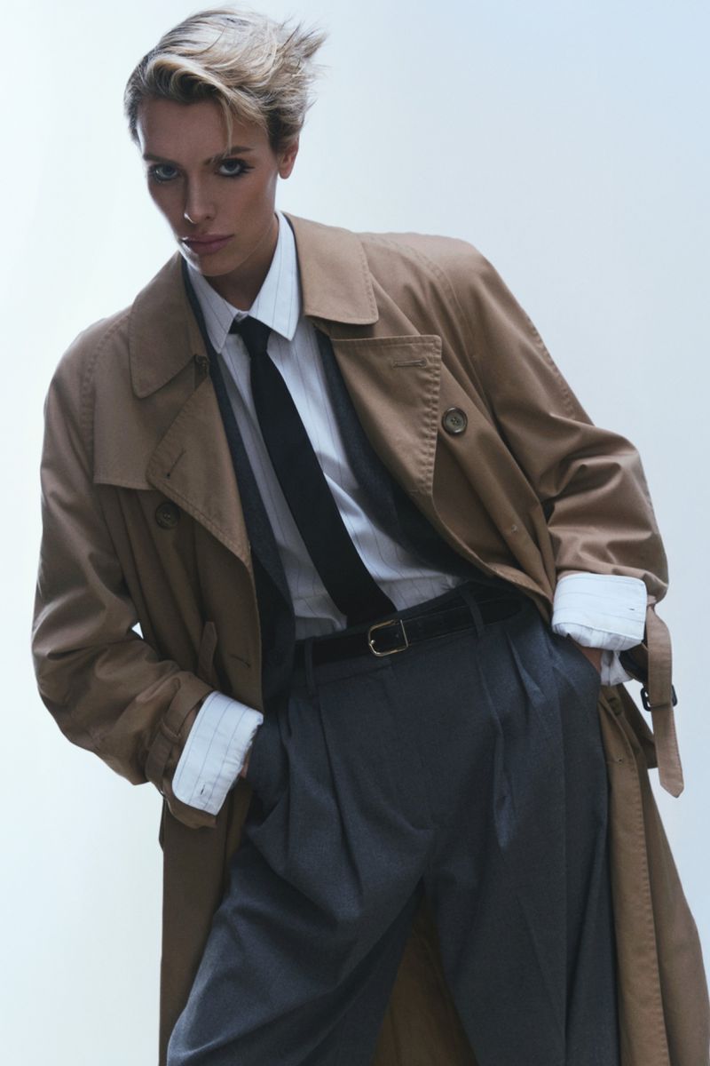 An adult with a short blonde haircut confidently poses wearing a beige trench coat over a collared shirt and tie against a light background.