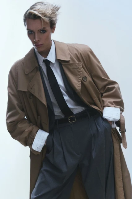 An adult with a short blonde haircut confidently poses wearing a beige trench coat over a collared shirt and tie against a light background.