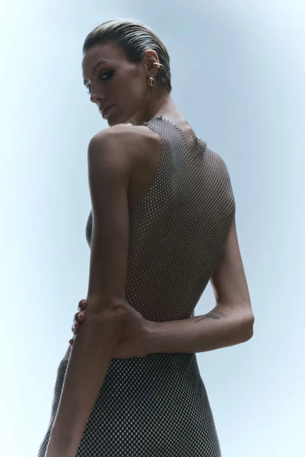 Woman with wet slicked-back hair, wearing silver halter dress, looking over shoulder in studio shot with bright lighting and pale blue backdrop.