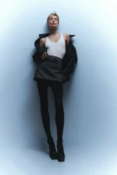 Wallis Day leaning against a blue studio wall wearing a white tank top, grey miniskirt, and black jacket.