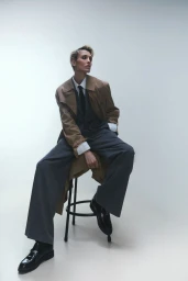 Wallis Day seated on a stool wearing an oversized camel trench coat, grey trousers, and a tie against a white seamless background