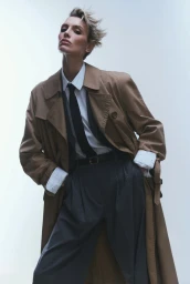 Wallis Day in a camel trench coat, white shirt, and black tie against a white background for Numero Netherlands editorial.