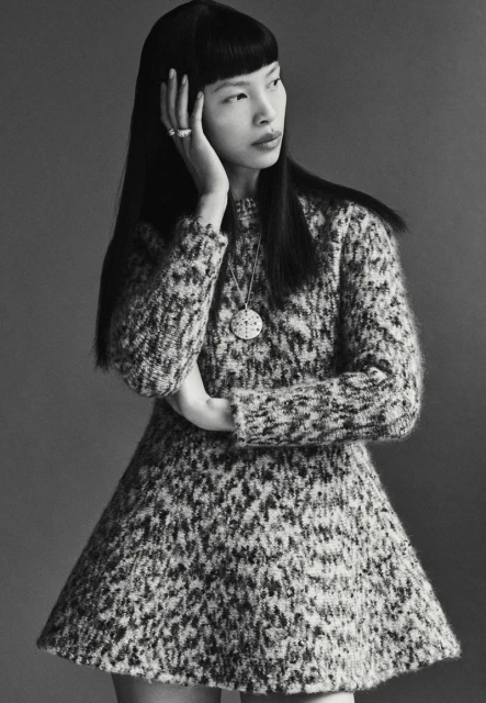 Monochromatic portrait of a stylish Asian woman in a knit dress, accented with a pendant necklace, rings, and a sleek hairstyle.