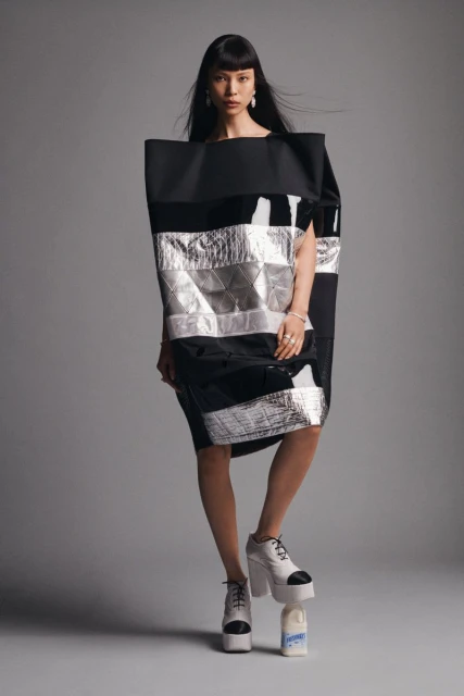 A stylish woman poses in a black and silver dress with platform shoes in a studio setting, fashion-forward with a unique outfit and makeup.
