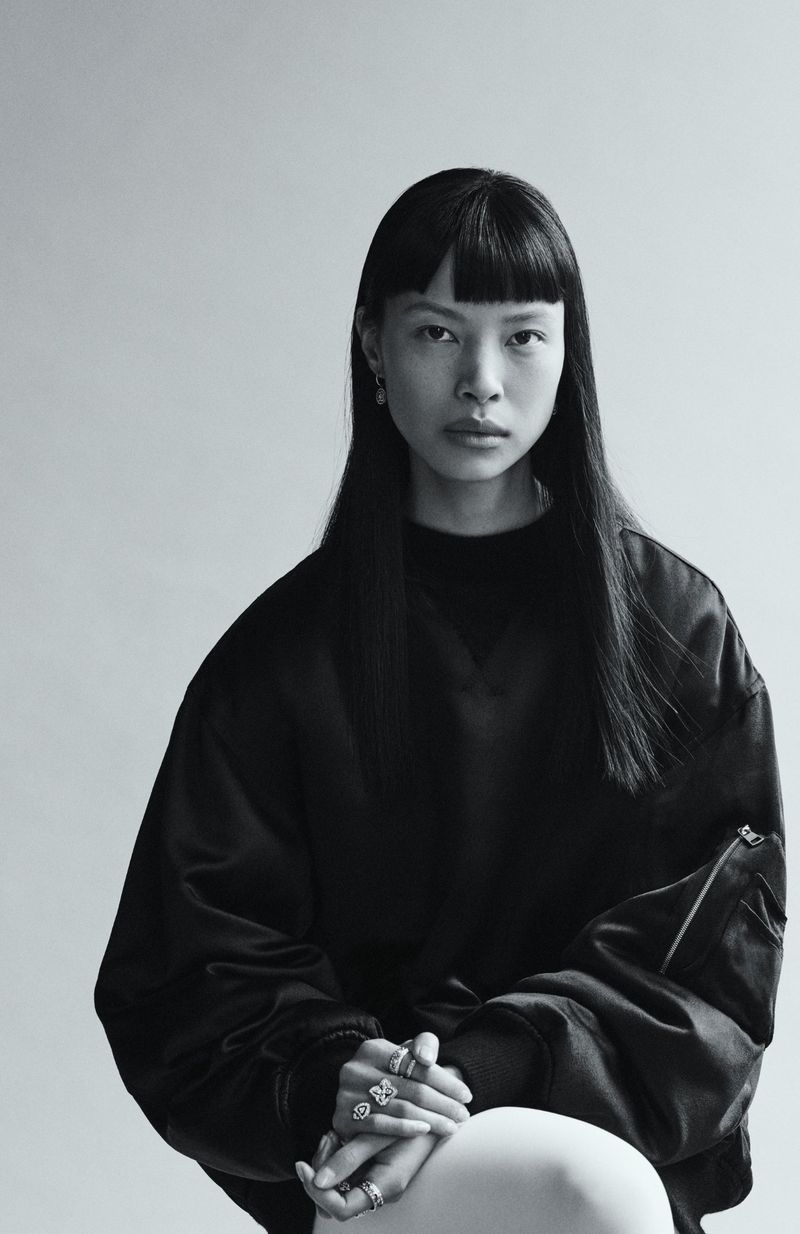 Model with long dark hair wearing a black bomber jacket against a neutral grey studio background