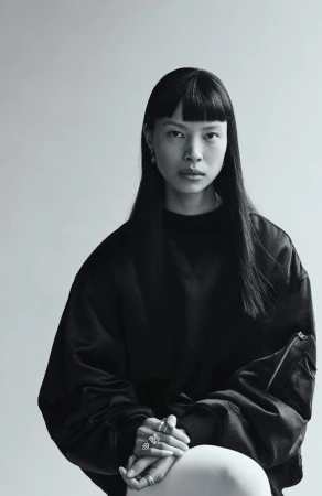 Model with long dark hair wearing a black bomber jacket against a neutral grey studio background