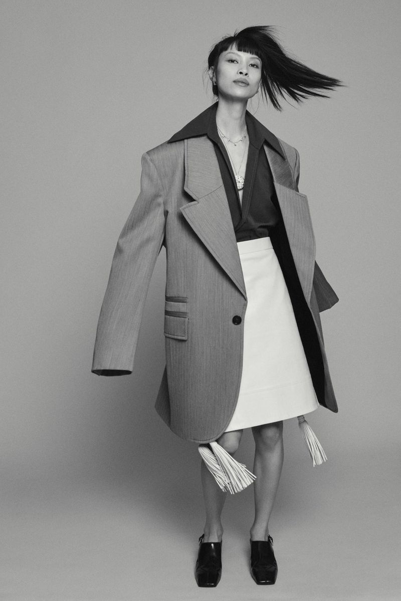 Black and white studio shot of an Asian woman in an oversized blazer and skirt, hair blowing, minimalist background.