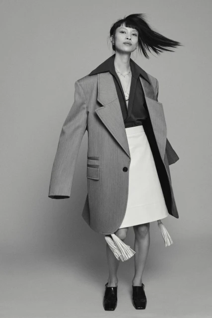 Black and white studio shot of an Asian woman in an oversized blazer and skirt, hair blowing, minimalist background.