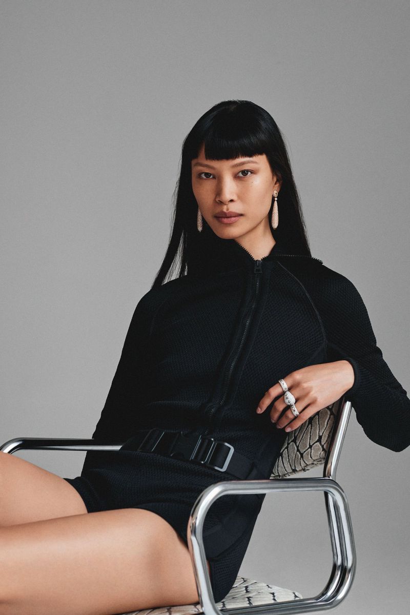 Portrait of a woman with black hair in black romper sitting in a metal chair against a grey backdrop in a studio setting.