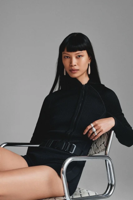 Portrait of a woman with black hair in black romper sitting in a metal chair against a grey backdrop in a studio setting.