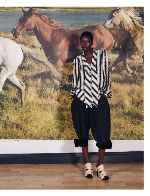 Editorial fashion shot of a Black woman in a striped blouse and black culottes posing indoors against a backdrop of horses running.