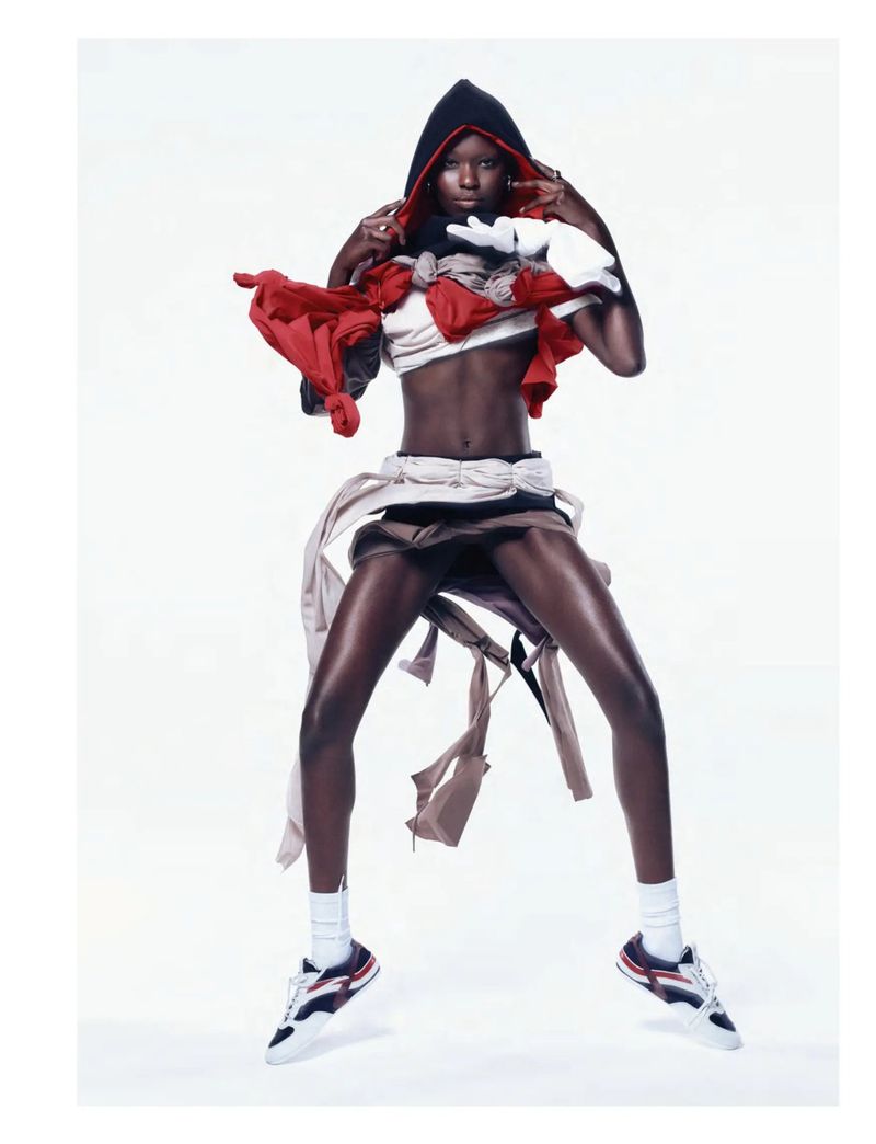 Fashionable African woman in athletic clothes and a black hood, jumping in a brightly lit white studio. Red, white, and beige materials accent the outfit.