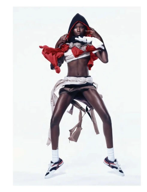 Fashionable African woman in athletic clothes and a black hood, jumping in a brightly lit white studio. Red, white, and beige materials accent the outfit.