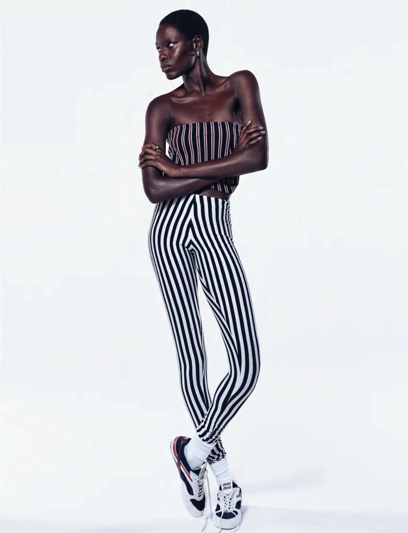 Full-length portrait of a slender, dark-skinned Black woman with short hair, wearing striped pants and top, posing in a photo studio.