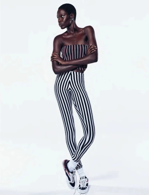 Full-length portrait of a slender, dark-skinned Black woman with short hair, wearing striped pants and top, posing in a photo studio.