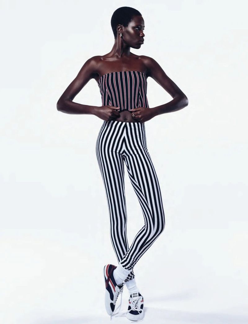 Fashion shot of a slender Black woman wearing a striped top and leggings, posing against a white backdrop with sneakers and white socks.