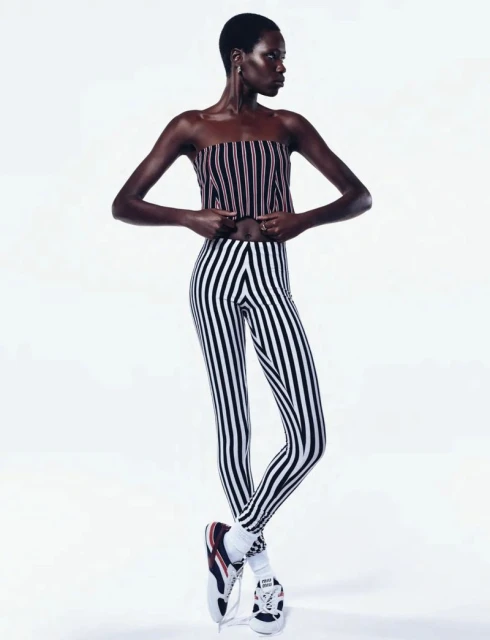 Fashion shot of a slender Black woman wearing a striped top and leggings, posing against a white backdrop with sneakers and white socks.
