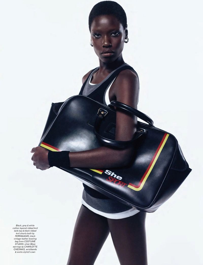 A stylish dark-skinned woman with short hair poses indoors holding a large black leather duffel bag with red and yellow stripes.