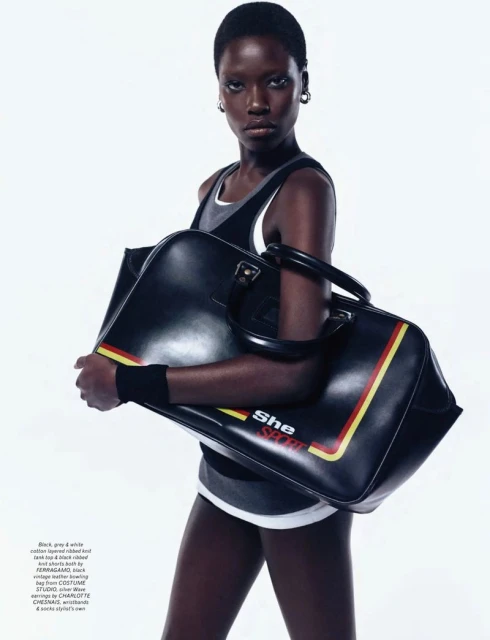 A stylish dark-skinned woman with short hair poses indoors holding a large black leather duffel bag with red and yellow stripes.