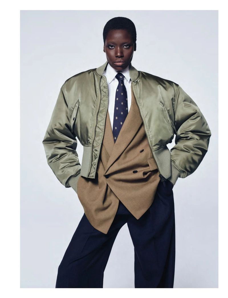 Fashion shot of an African female model posing in an olive bomber jacket, tan blazer, white shirt, and dark tie against a white background.