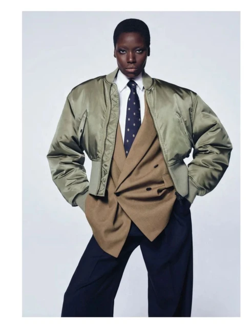 Fashion shot of an African female model posing in an olive bomber jacket, tan blazer, white shirt, and dark tie against a white background.