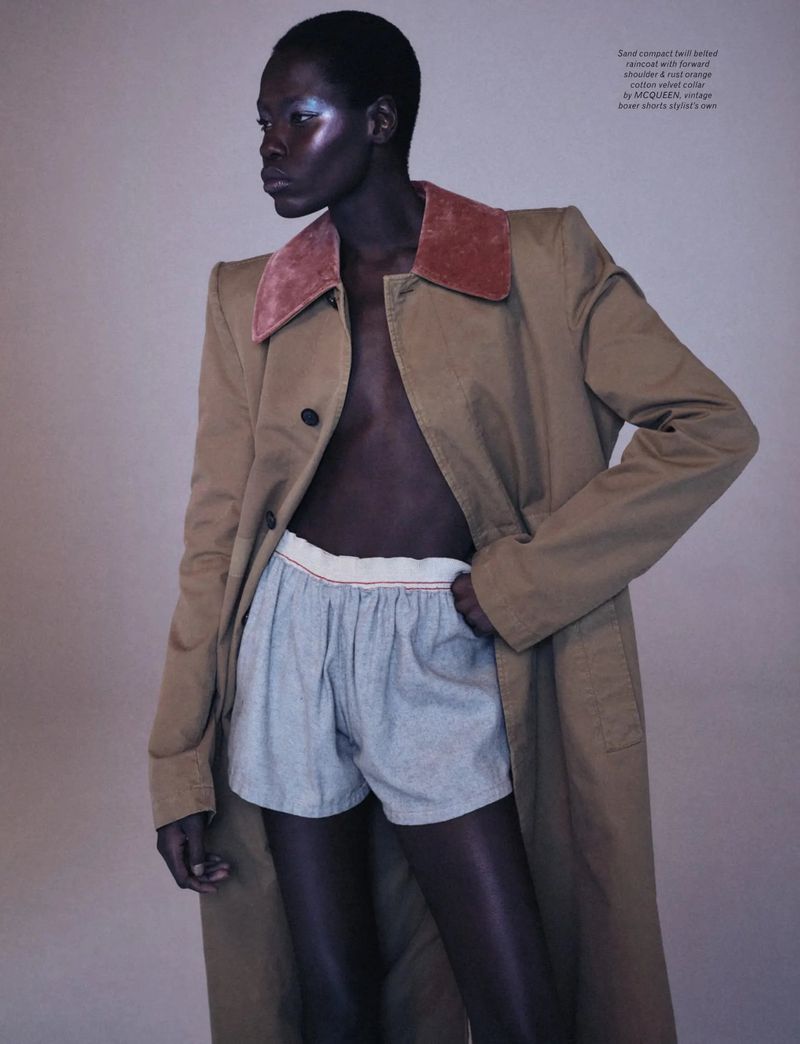 Fashion shot of a model wearing a tan trench coat and shorts, with a light background in a photo studio, looking away from the camera.