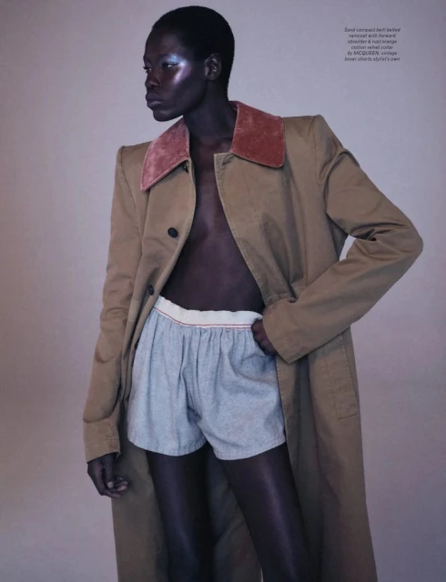 Fashion shot of a model wearing a tan trench coat and shorts, with a light background in a photo studio, looking away from the camera.