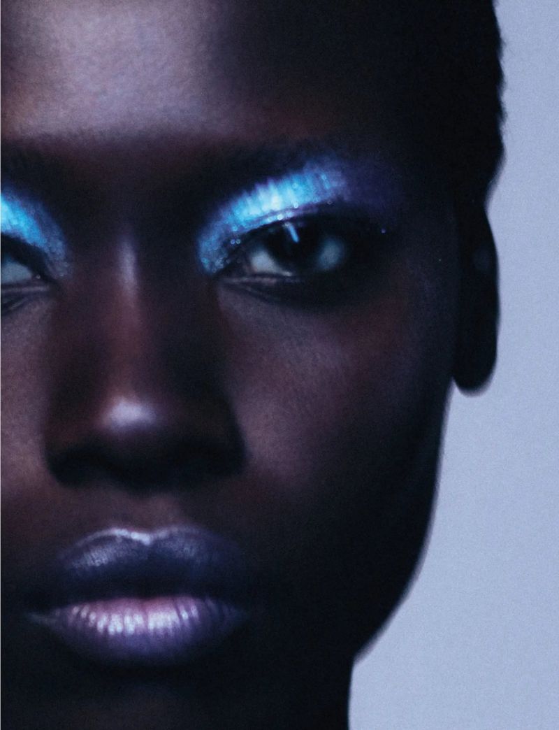 Close-up portrait of an African American woman with short hair, bold blue eyeshadow, and shimmering lipstick, against a light background.