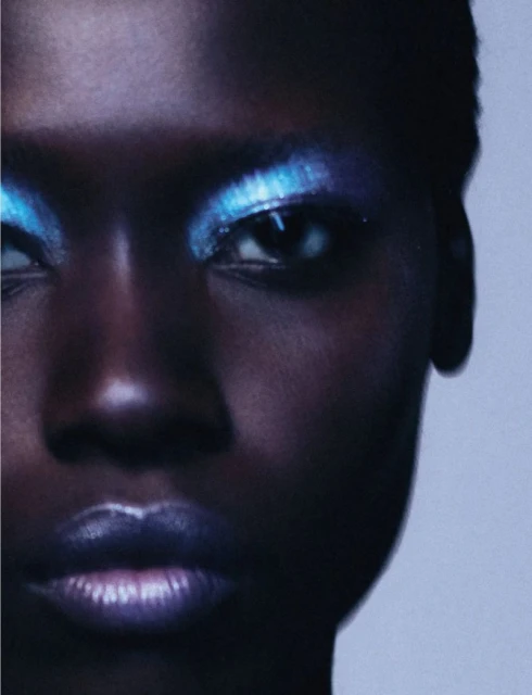 Close-up portrait of an African American woman with short hair, bold blue eyeshadow, and shimmering lipstick, against a light background.
