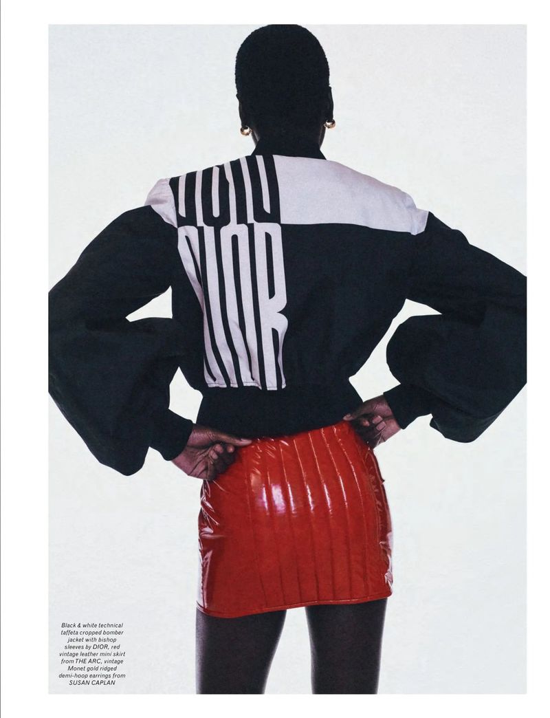 A Black model poses with their back to the camera, wearing a black and white jacket and a red mini skirt in a studio environment.
