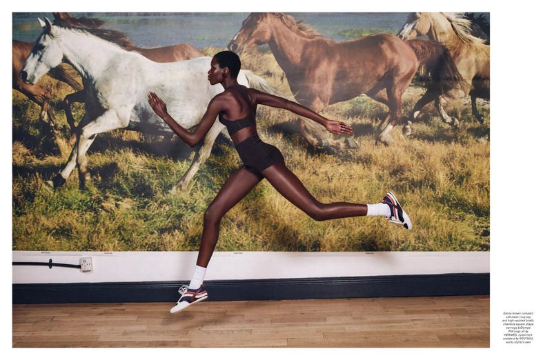 Athletic dark-skinned woman in black workout clothes running in front of a painted mural of wild horses in an indoor setting.