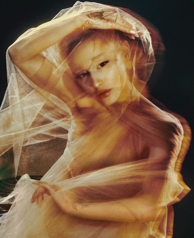 Woman with light skin poses with a sheer fabric draped around her head and body in a studio setting, showcasing soft lighting and artistic style.