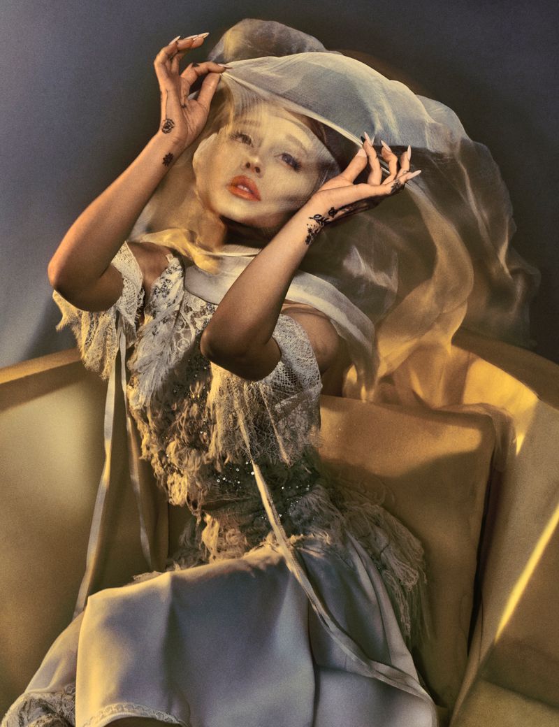 Woman in an ornate dress and sheer veil sits on a cream sofa in a warm, filtered light. She has her hands near her face.