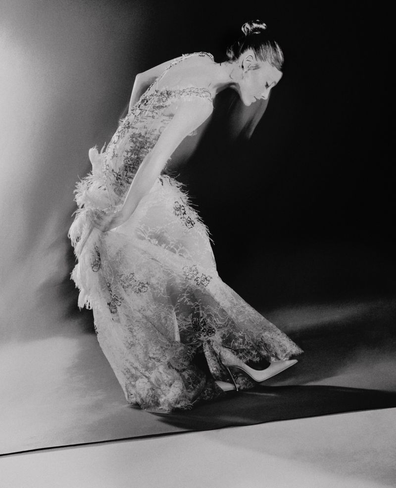 A full-length black and white portrait of a woman in a lace gown with feather accents, captured in motion against a dark background.