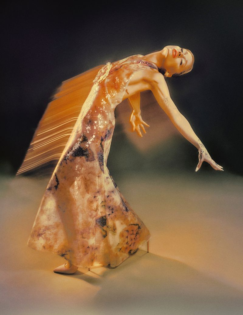 Elegant adult female in a flowing gown arches backward in a motion blur, set against a dark studio background with soft lighting.