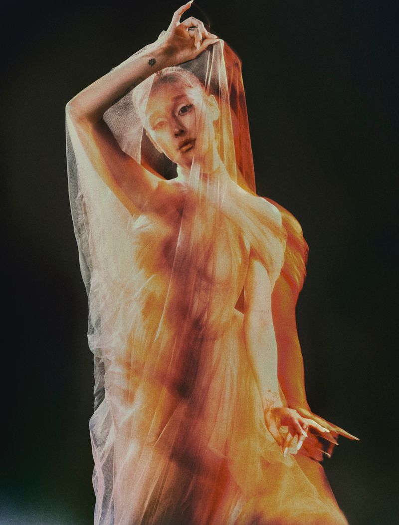 Artistic studio shot of a pale-skinned woman draped in sheer fabric, hands raised against a dark background with warm, diffused lighting.