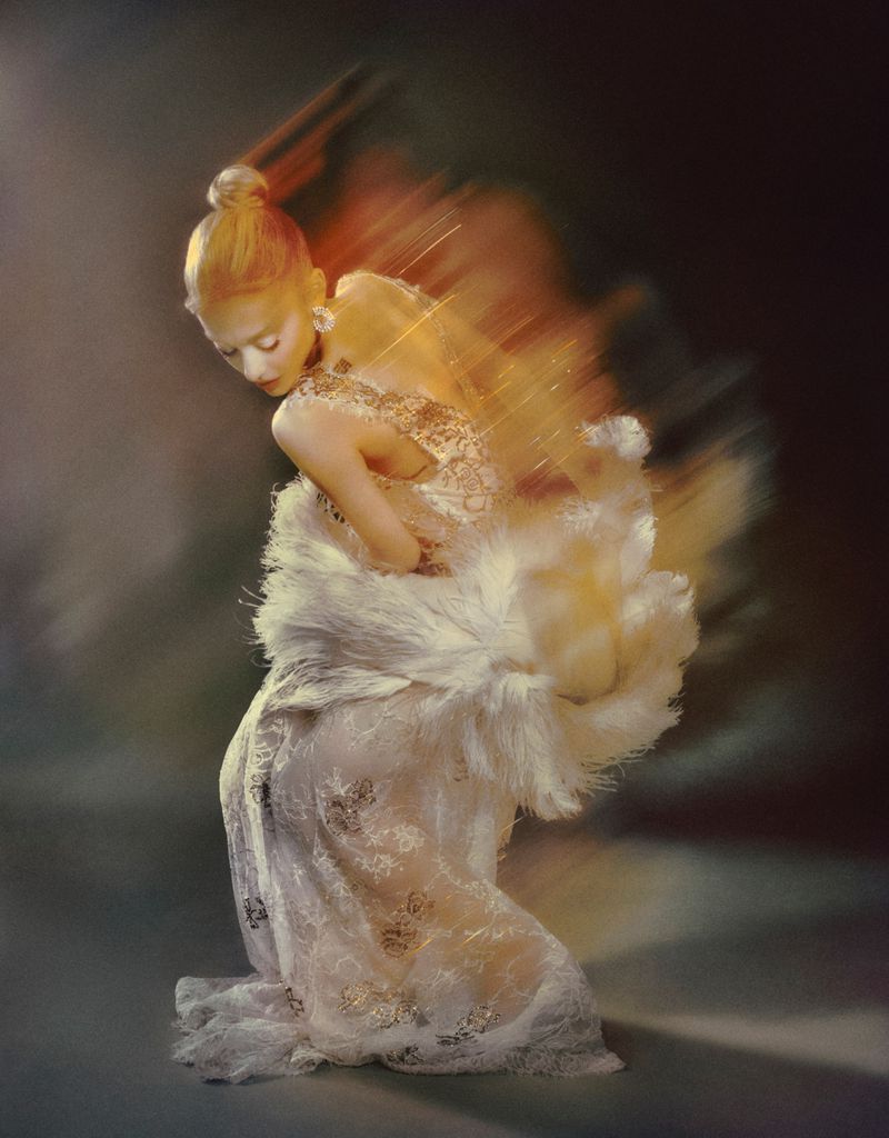 Artistic fashion image of a woman with fair skin and auburn hair wearing a white lace dress and feathered boa, set against a gray background.