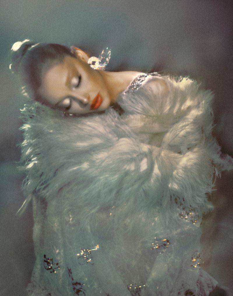 Elegant woman with closed eyes in a feathered white dress and crystal earrings against a softly blurred background.