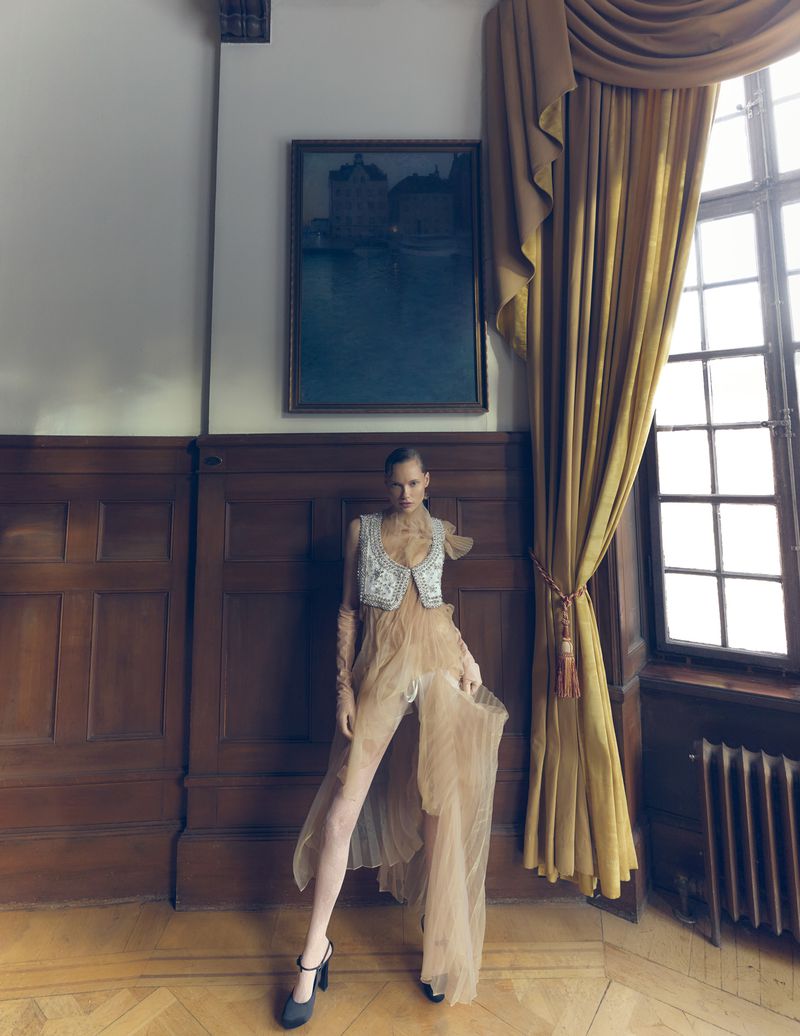 Woman poses in flowing gown and heels near window and panelled wall, framed by golden curtains, against a backdrop of art and interior details.