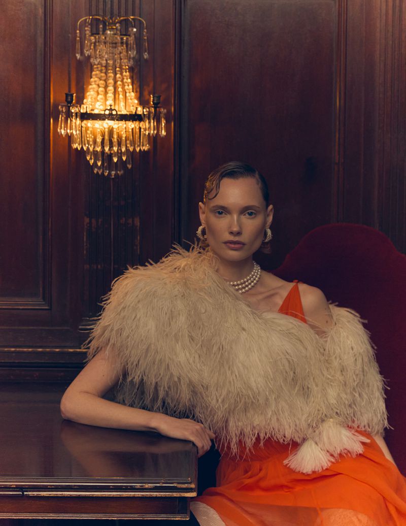 Glamorous woman poses in an orange dress and feathered wrap under a chandelier. The scene evokes an elegant vintage aesthetic.