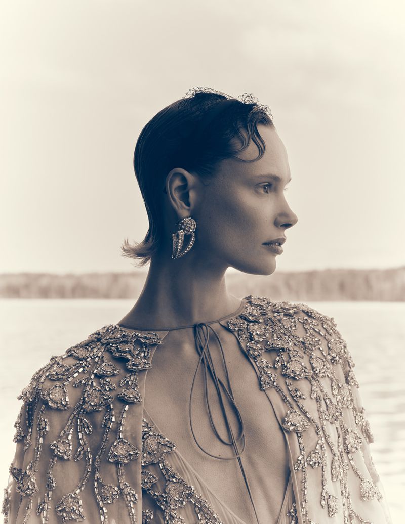 A woman in an embellished cape poses in front of a body of water, showcasing fashion, beauty, and an elegant profile.