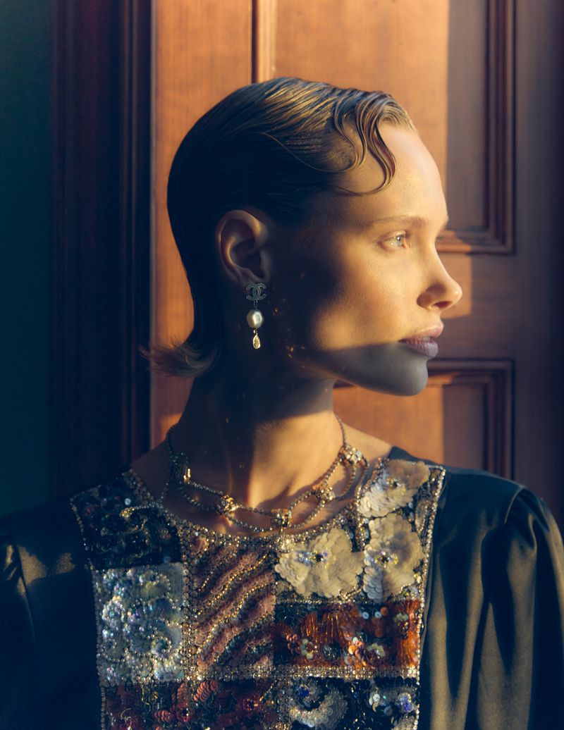 Profile shot of a woman with short hair wearing statement earrings and an embellished top in front of a paneled door with warm lighting.