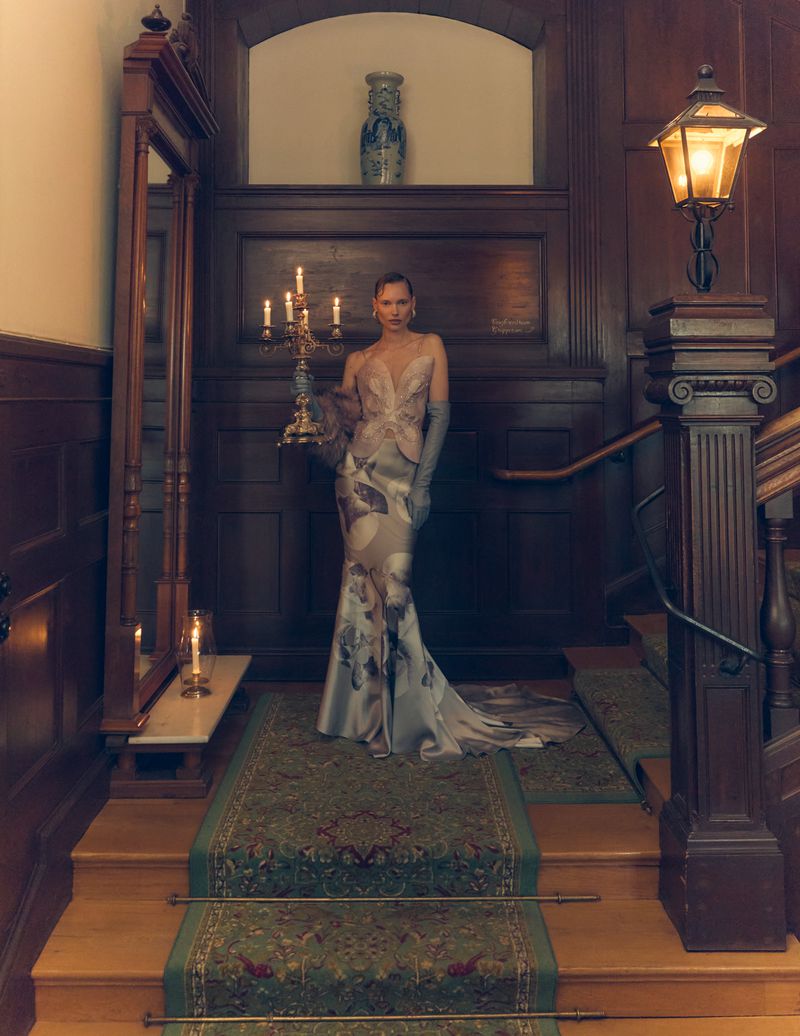 Woman in a formal gown holding a candelabra on a staircase inside an elegant home with wood paneling and dark lighting for fashion editorial.