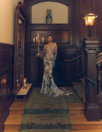 Woman in a formal gown holding a candelabra on a staircase inside an elegant home with wood paneling and dark lighting for fashion editorial.