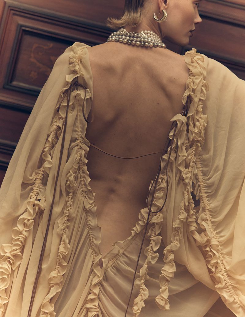Back view of a woman in a light yellow gown adorned with ruffles, wearing a pearl necklace and earrings against a dark wood background.