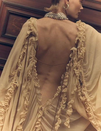 Back view of a woman in a light yellow gown adorned with ruffles, wearing a pearl necklace and earrings against a dark wood background.
