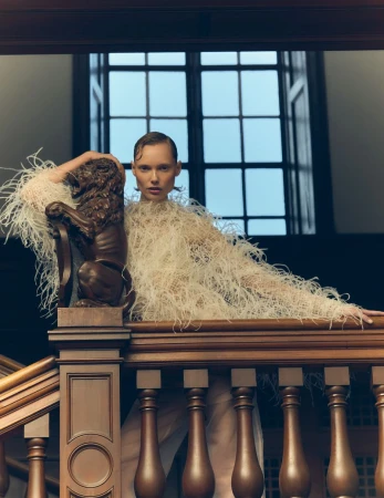 Woman in feather dress poses on a wooden staircase with a decorative lion statue in an indoor setting with natural window light.